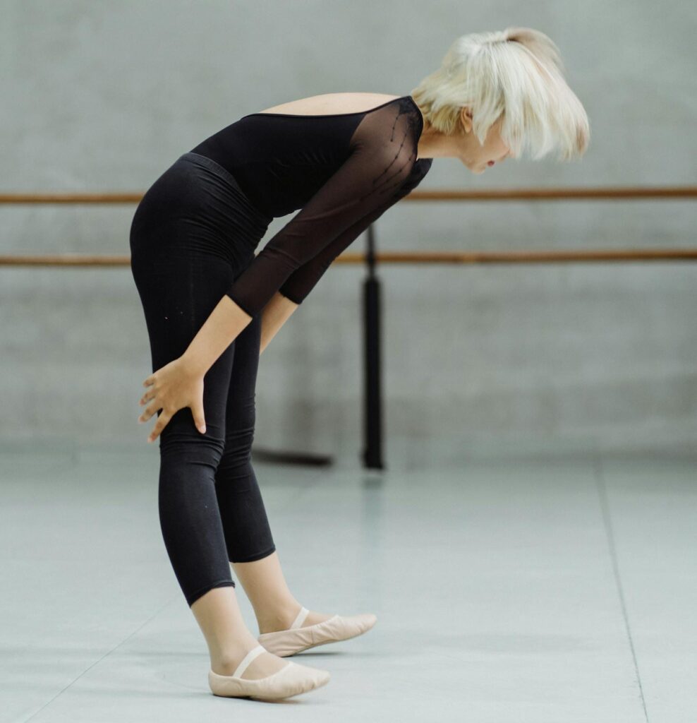 A female ballet dancer in a black leotard stretches in a studio, showcasing flexibility and grace.
